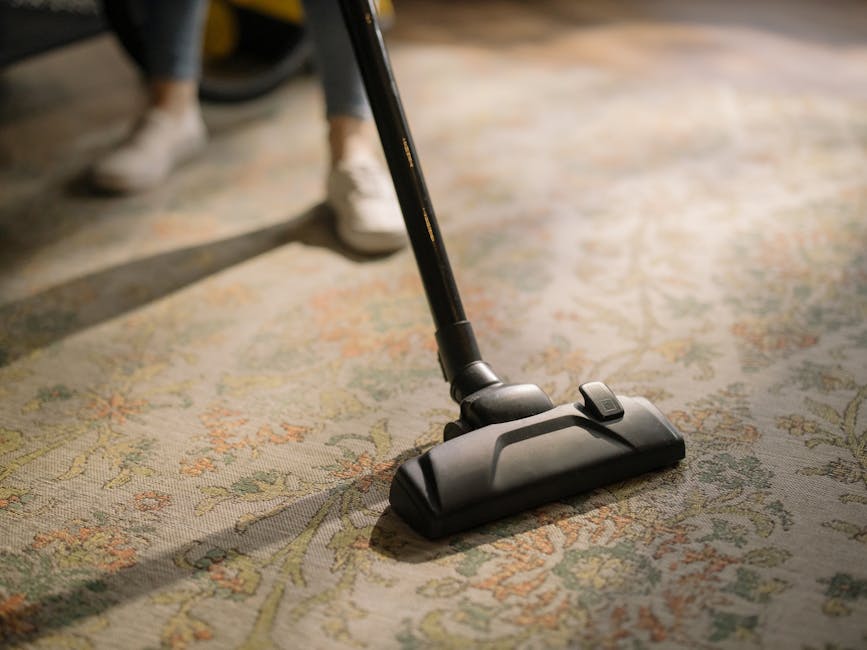 A woman in a casual olive green jacket, dark jeans, and white sneakers is performing deep carpet cleaning in a living room. She is using a yellow and black vacuum cleaner with an attached hose and a n