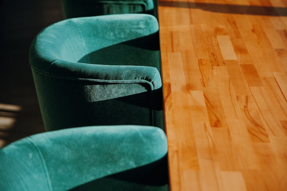 Close-up of a person's hand using a handheld vacuum cleaner to deep clean the light green upholstered chair in a living room setting, with a dark gray sofa in the background. The cushions of the armch